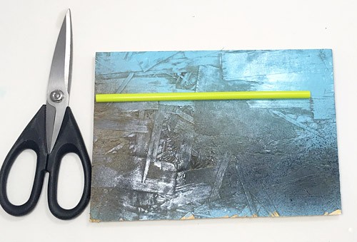 A straw lays on a wooden board next to a pair of scissors