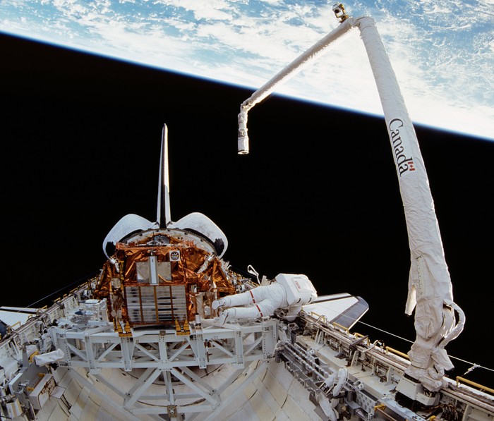 A picture taken in space of a robotic arm assisting an astronaut on the exterior of the space shuttle, with the Earth in the background