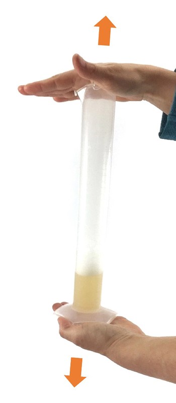 Two hands holding a measuring cylinder filled with liquid. Arrows point up and down from the cylinder.