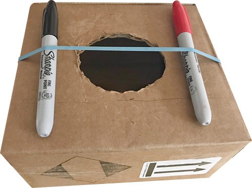 A rubber band secures two sharpies on either side of a hole cut into a cardboard box