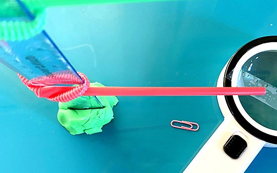 Top down view of a ruler held straight up by a lump of clay. A knot in a straw holds the straw to the ruler so a long piece of it sticks out horizontally.