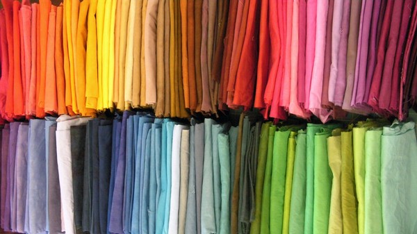 Two rows of dozens of fabrics dyed in many solid shades of red, orange, yellow, brown, blue, green, and purple.