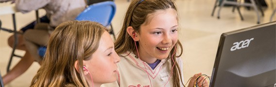 Two students with earbuds sit in front of a computer