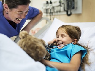 A nurse assistant smiles at a young girl in a hospital bed who is listening to a teddy bear’s chest with a stethoscope.