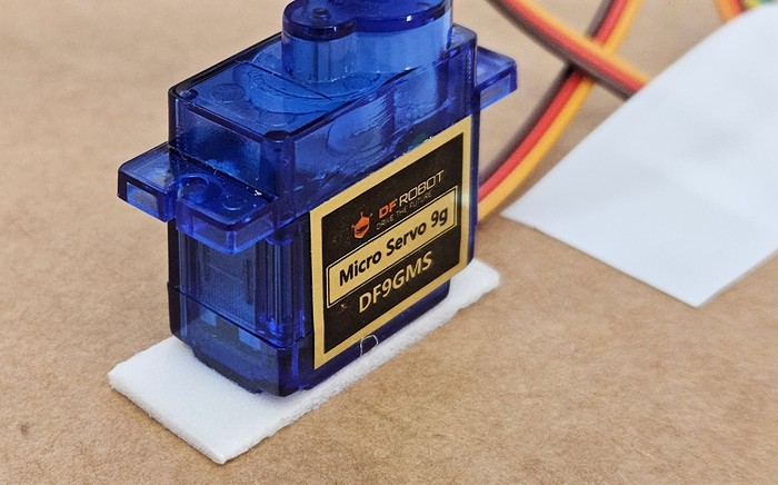 Micro servo motor with sitting upright on a piece of double-sided foam tape which is on top of a piece of cardboard