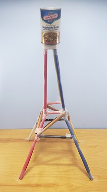 A paper tower with a truss structure that is wider at the bottom and narrow at the top, with a can of beans at the top.