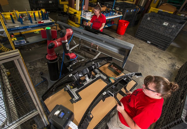 A woman at a table holds a mechanical part while a robotic arm performs work next to her. 