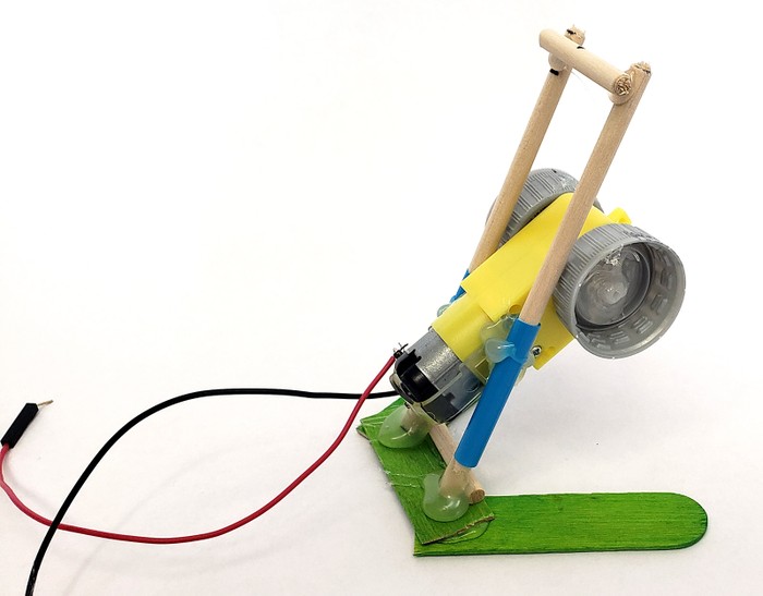 Wooden dowels supporting the motor are glued upright at a steep angle to attach to the base of the robot. A crosspiece at the top connects the two dowels.