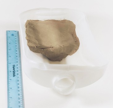 Front view: A gallon jug cut in half, lengthwise, in which a clay platform is formed in the area farthest away from the spout. A ruler indicates the pavement is about 10 cm long.