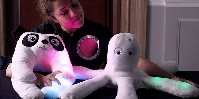 A woman sits next to two robots, a dog and an octopus, that are soft and cute. They are covered in fabric and stuffing, like plush toys.