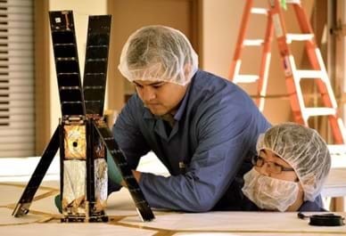 Two men wearing hair and beard nets examine a device on a table. Four solar panel-covered sides of the tall rectangular object (shoe-box sized) are flipped up or away to reveal the inside volume of the satellite cube, which contains a folded-up silver and gold fabric.