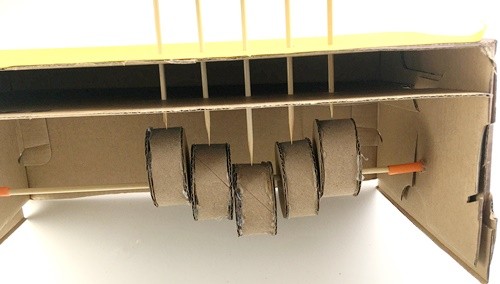 5 parallel skewers piercing through the top of a box and a second piece of cardboard. Each skewer rests on one of five driving cylinders.