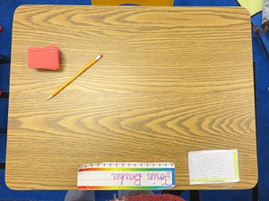 Overhead view of a stack of paper squares, pencil, notecard and nametag on a small wooden desk