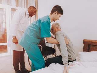 A nurse assistant helps to lift an older man off a bed and helps him into a wheelchair.