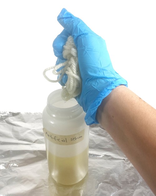 A hand holding a piece of wool yarn above a bottle filled with yellow liquid.