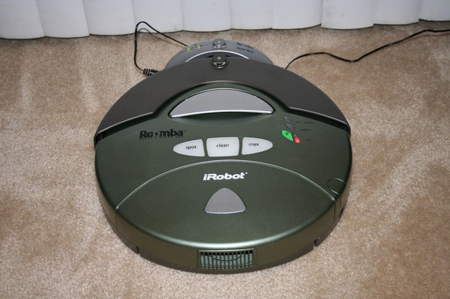 A round robotic vacuum cleaner about the diameter of a dinner plate and roughly two inches tall docked at its charging station. 
