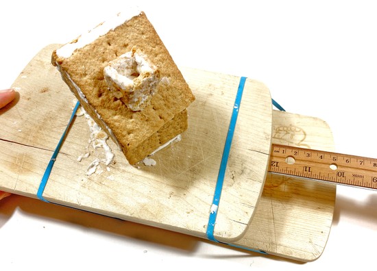 A house made from Graham crackers is standing on a self-made shake table made of ping pong balls and two cutting boards. The top plate of the shake table is pulled 5 centimeters to the side. 