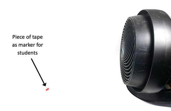 A fan is standing on a table. A piece of tape is located on the table about 12 inches away from the fan.