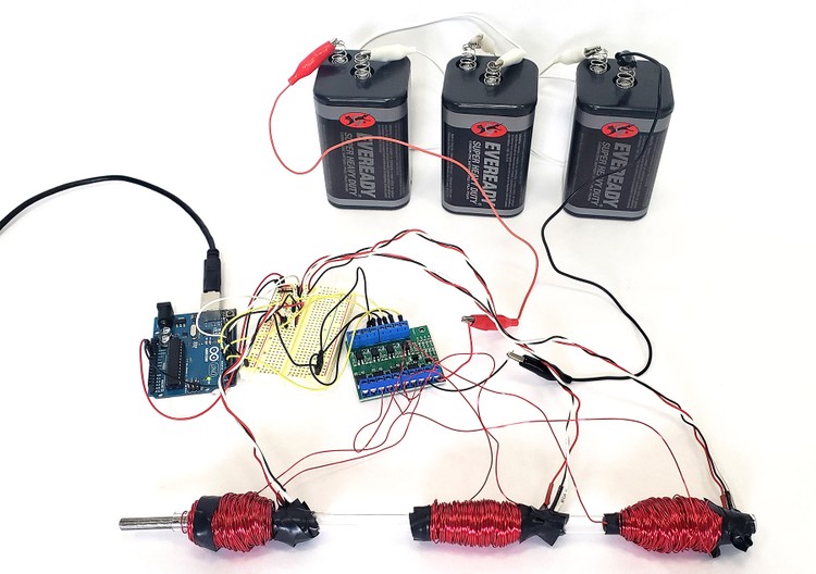 An Arduino, a circuit board, three lantern batteries, and a clear plastic tube with three coils of wire around it.