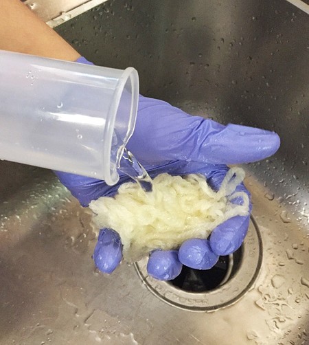 A gloved hand holding a piece of wool yarn in the sink. Water is poured over the wool from a measuring cylinder.