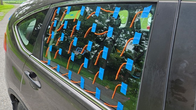 The window of a car is covered with evenly spaced pieces of painter's tape. Each piece of tape secures one end of a short strand of yarn.