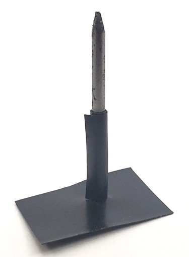 A nail standing vertically on its head, with one flat piece of electrical tape at the bottom and another piece wrapped around the base