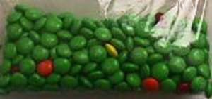 Clear bag full of green M&Ms with one yellow and five red M&Ms interspersed.