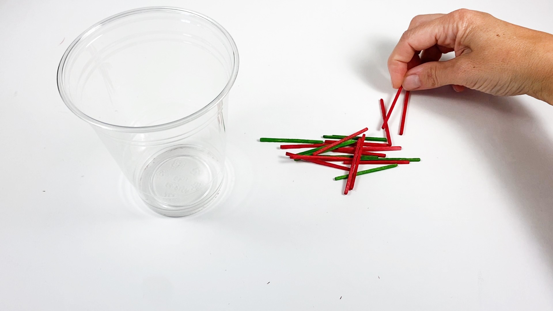 A pile of green and red matchsticks next to a plastic cup. A hand is holding one of the red matchsticks.